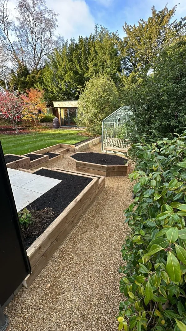 A professionally designed garden featuring tiered wooden sleeper beds, a hexagonal planter, a greenhouse, and a modern garden room.