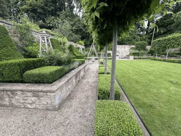 A gravel pathway lining a formal garden, bordered by stone walls, low box hedges, and tall pleached trees.
