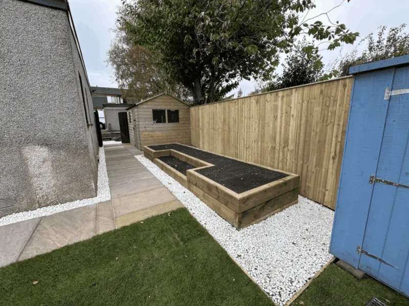 Raised wooden sleeper flower beds constructed in a narrow side garden alongside a white gravel path and wooden shed.