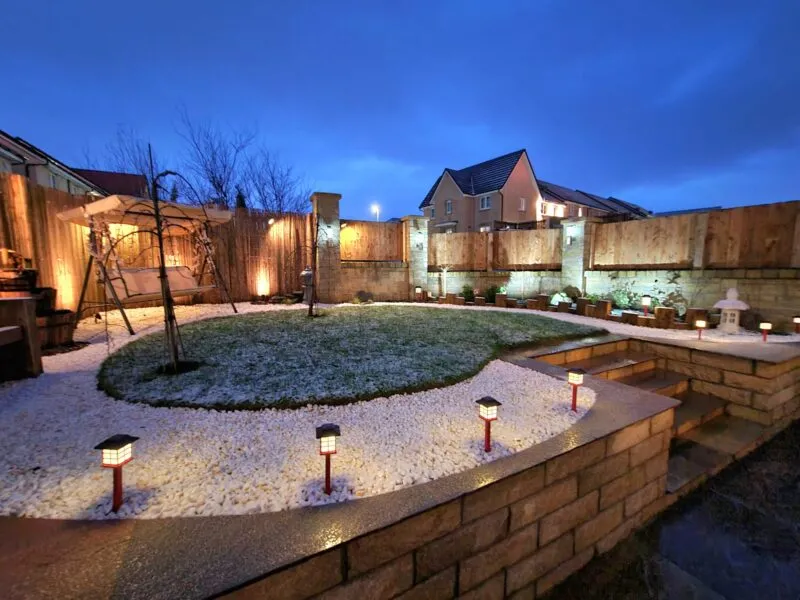 A landscaped garden illuminated at night, featuring bollard lighting, a circular lawn, white gravel, and a swing seat.