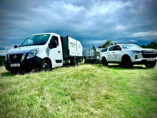A Stephen Paul Gardens tipper van and Isuzu pickup truck parked in a grassy field under a dramatic cloudy sky.