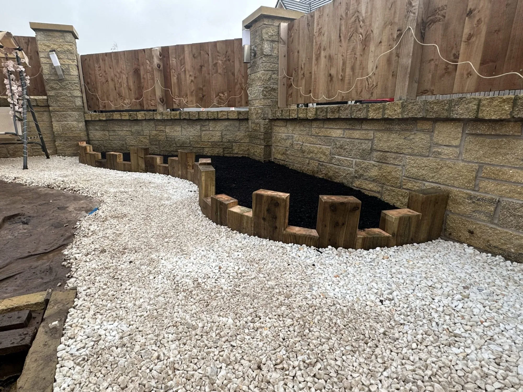 A curved garden border constructed with vertical wooden sleepers alongside a decorative white gravel path and stone wall.