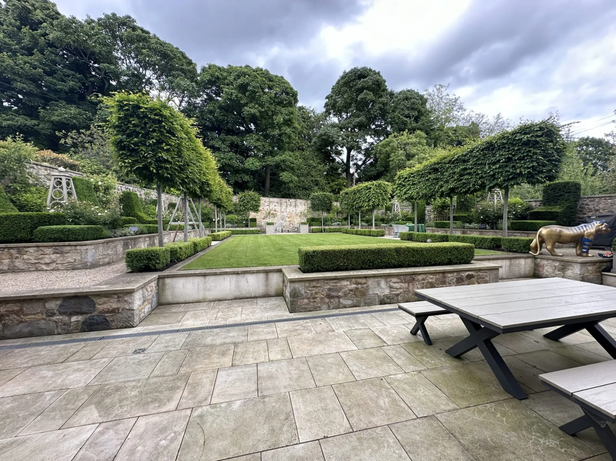 A high-end formal garden design featuring a spacious stone patio, manicured lawn, pleached trees, and modern outdoor furniture.