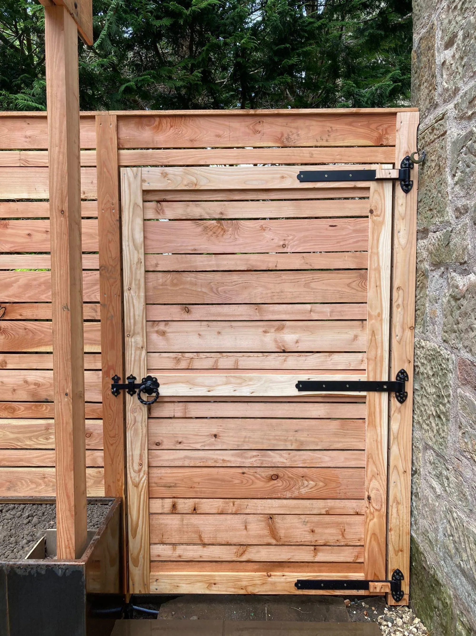 A bespoke wooden garden gate featuring horizontal slats and black iron hardware, installed next to a stone wall.