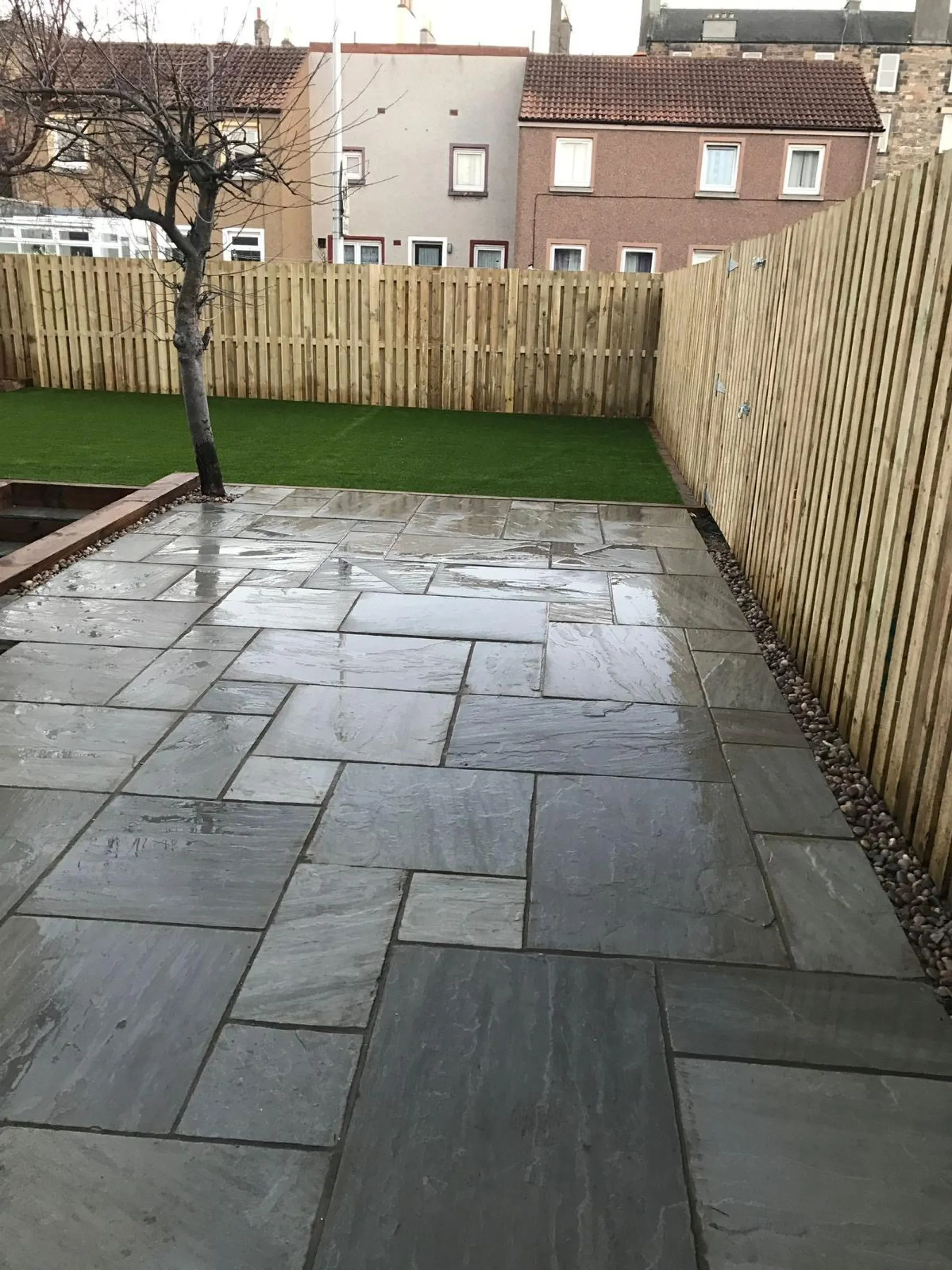 Photo of a grey natural stone patio paving installation in an enclosed garden with a timber fence and artificial lawn by Stephen Paul Gardens.