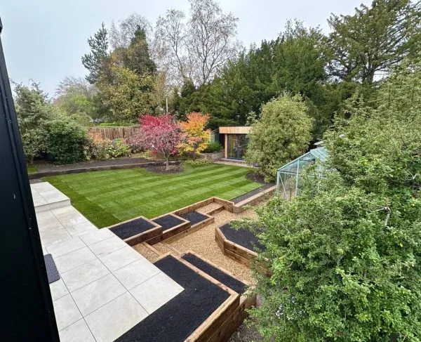 A completed garden landscaping project featuring a pristine striped lawn, tiered wooden sleeper flower beds, a greenhouse, and a modern garden room.