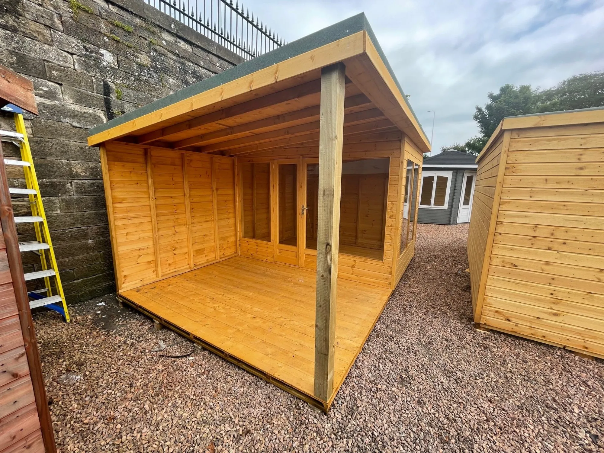 A timber garden room or summerhouse featuring a covered veranda and glass doors, displayed on a gravel surface.