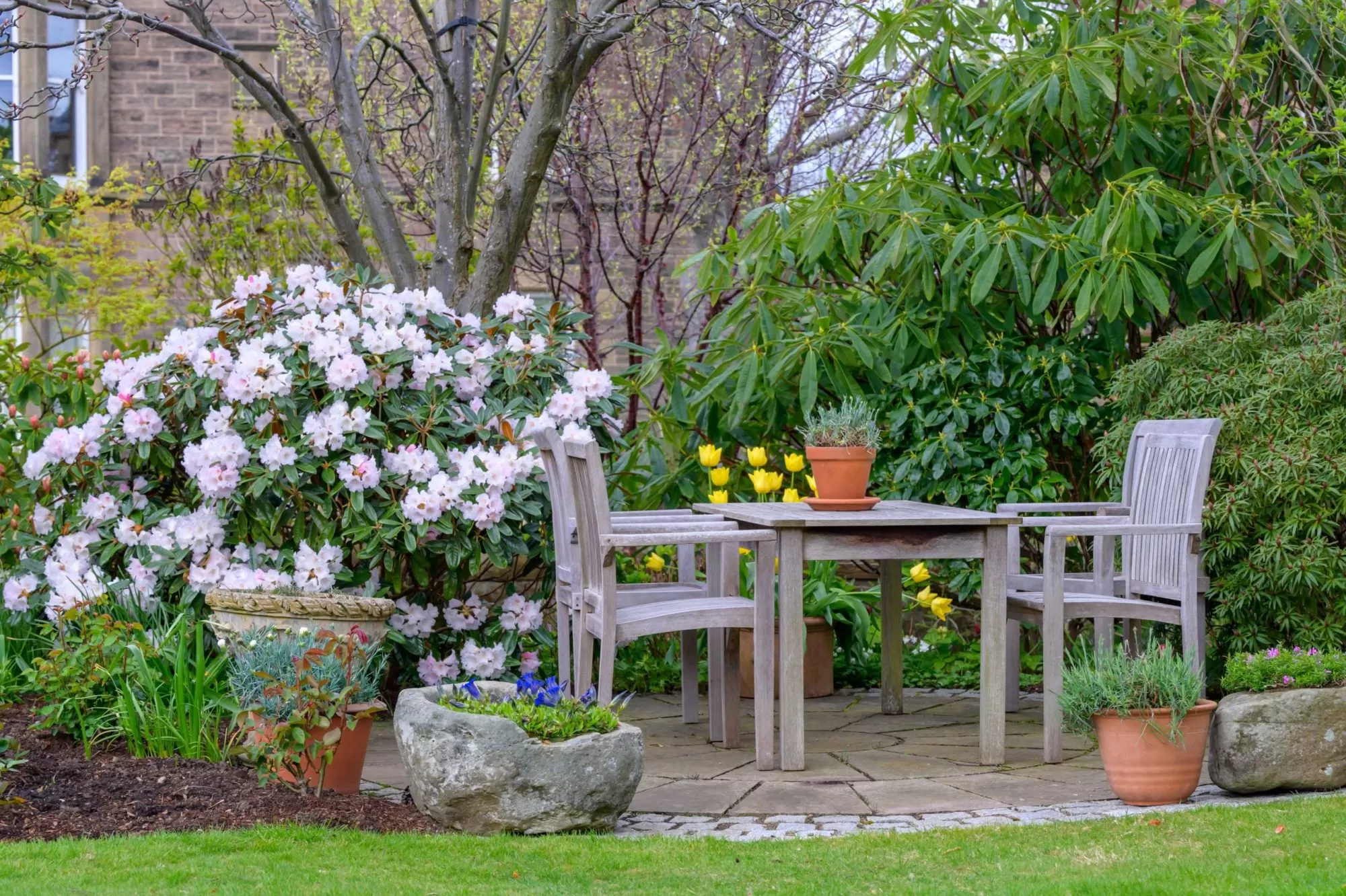 Photo of a wooden table and chairs on a circular stone patio surrounded by blooming rhododendrons by Stephen Paul Gardens.